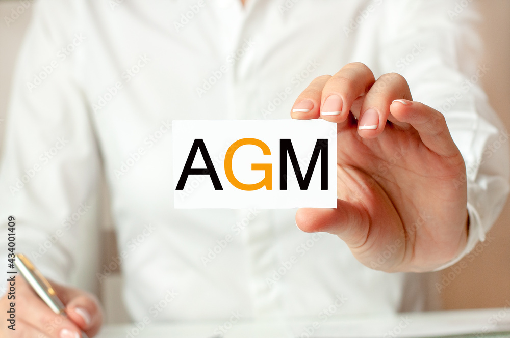 A woman in a white shirt holds a piece of paper with the text: AGM. AGM - annual general meeting. Business concept. A woman in a white shirt holds a piece of paper with the text: AGM. AGM - annual general meeting. Business concept.
