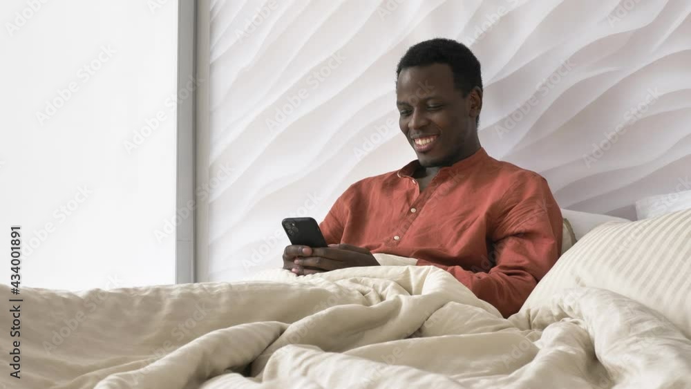 Handsome African-American man in pajamas laughs reading message on smartphone sitting on bed in sunny morning slow motion Handsome African-American man in pajamas laughs reading message on smartphone sitting on bed in sunny morning slow motion