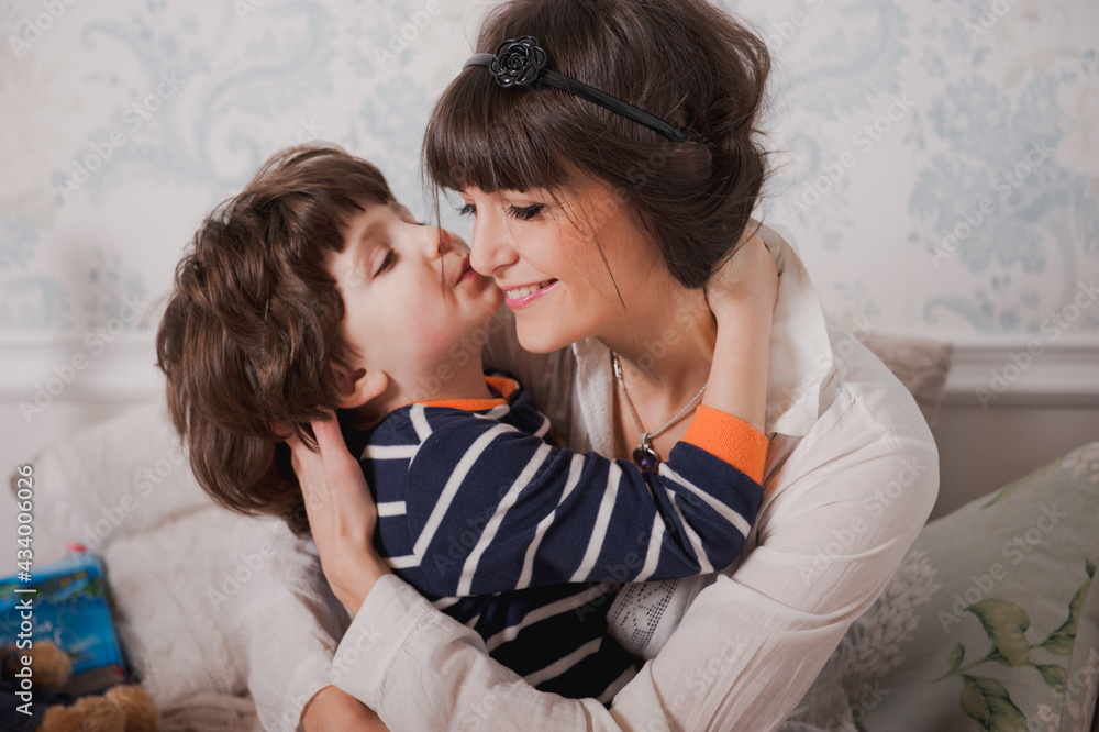 Boy kisses his mom and hugs. Happy young woman have fun with cute child baby boy. Mom and son sit on the bed before going to bed. Good night concept Boy kisses his mom and hugs. Happy young woman have fun with cute child baby boy. Mom and son sit on the bed before going to bed. Good night concept