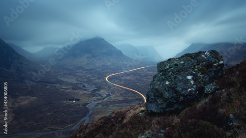 Morning landscape with car lights on the mountain road leading to Glencoe and a big rock in the foreground. A82 road opposite Buachaille Etive Mor before dawn. Scotland photo