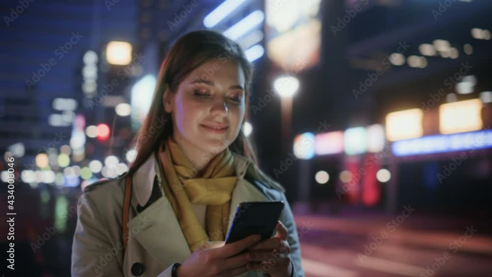 Portrait of a Beautiful Woman in Trench Coat Walking in a Modern City Street with Neon Lights at Night. Attractive Female Using Smartphone and Looking Around the Urban Cinematic Environment.