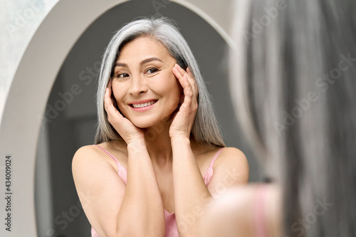Obraz na plátně Happy middle 50 years aged asian woman with gray hair looking at mirror reflection examining touching face enjoying antiaging beauty treatments
