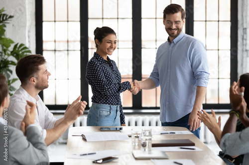 Happy Indian female team leader shaking hands with proud employee, thanking promoted worker for good job and congratulating with reward. Millennial business group welcoming newcomer with applause