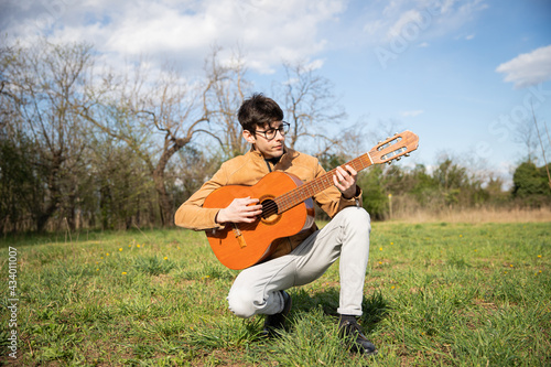 Young musician, kneeling plays the classical guitar in a field in the open air.