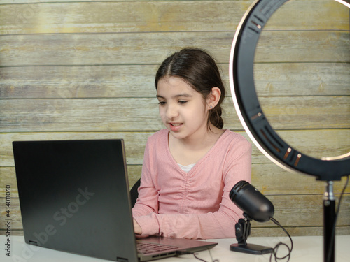 Small girl using camera of laptop in front of ring light, shooting video for blog. Adorable child learning new technology