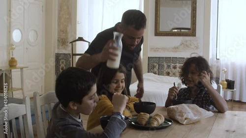 Solicitous Caucasian father pouring milk into kids plates. Cheerful children sitting at table, talking, eating and laughing. Fatherhood, food concept.