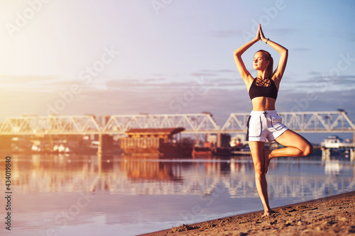 Woman practicing yoga meditation barefoot at sunrise on the sand. Morning yoga on the beach or coast of river on bridge urban city background