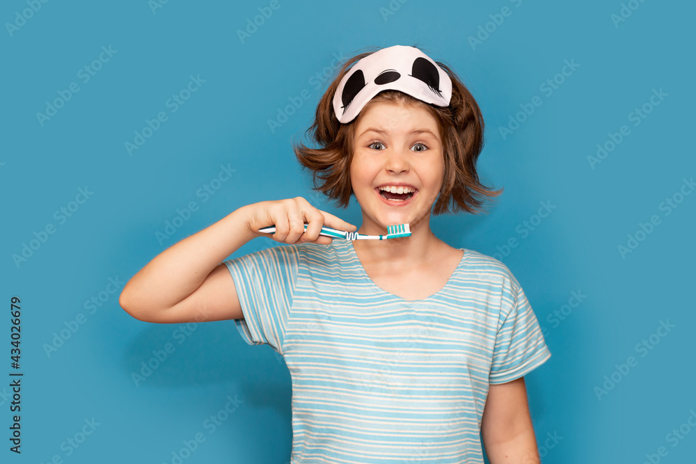 Fototapeta premium Happy child kid girl with sleeping mask brushing her teeth with toothbrush in morning isolated over blue background. Health care, dental hygiene. Mockup, copy space