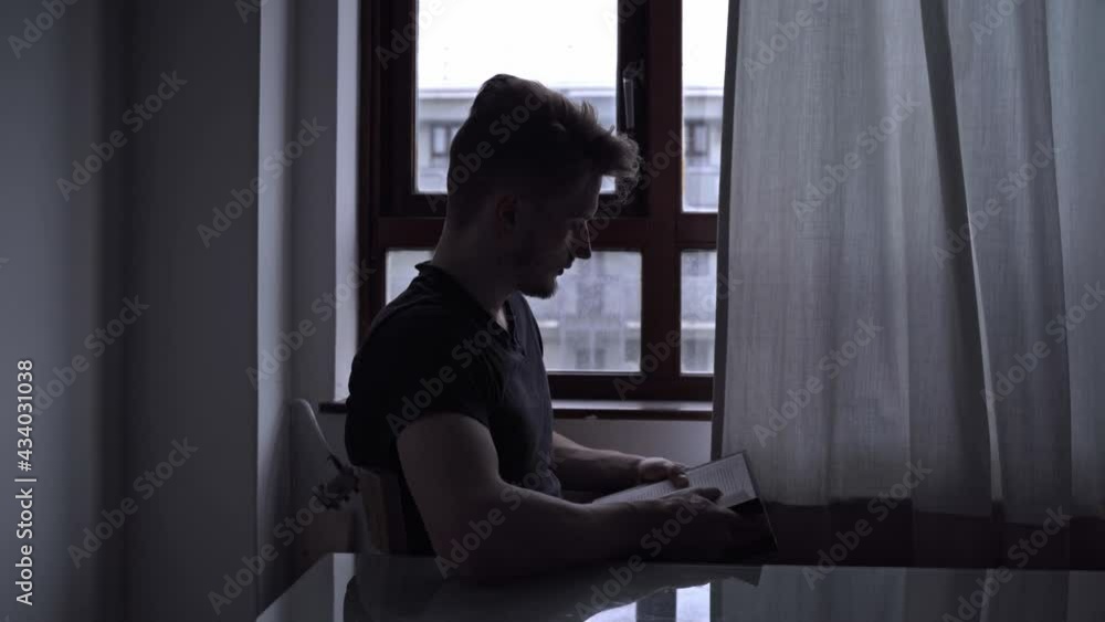 Young man sitting near a window in a dark room while reading and looking at the window on a cloudy rainy day. Young man sitting near a window in a dark room while reading and looking at the window on a cloudy rainy day.