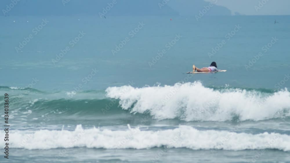 Slow motion shot of breaking surf waves and a surfer floating in the blue sea.