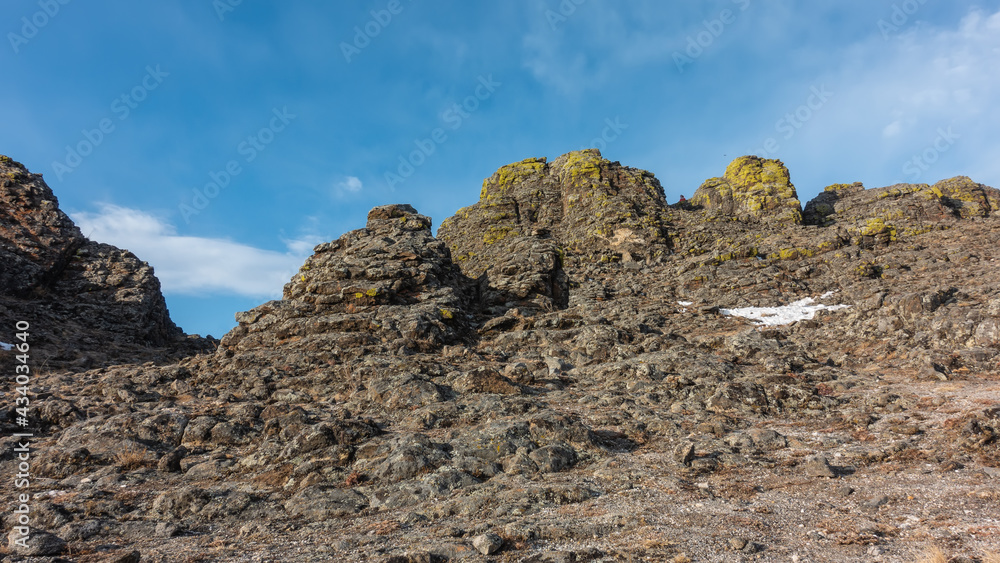 A picturesque mountain range against the backdrop of a bright blue sky. Yellow lichens grow on the stones, areas of snow lie.