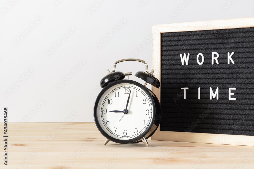 Classic black alarm clock on wooden table and black board with words 'work time' with white wall background and copy space. Classic black alarm clock on wooden table and black board with words 'work time' with white wall background and copy space.