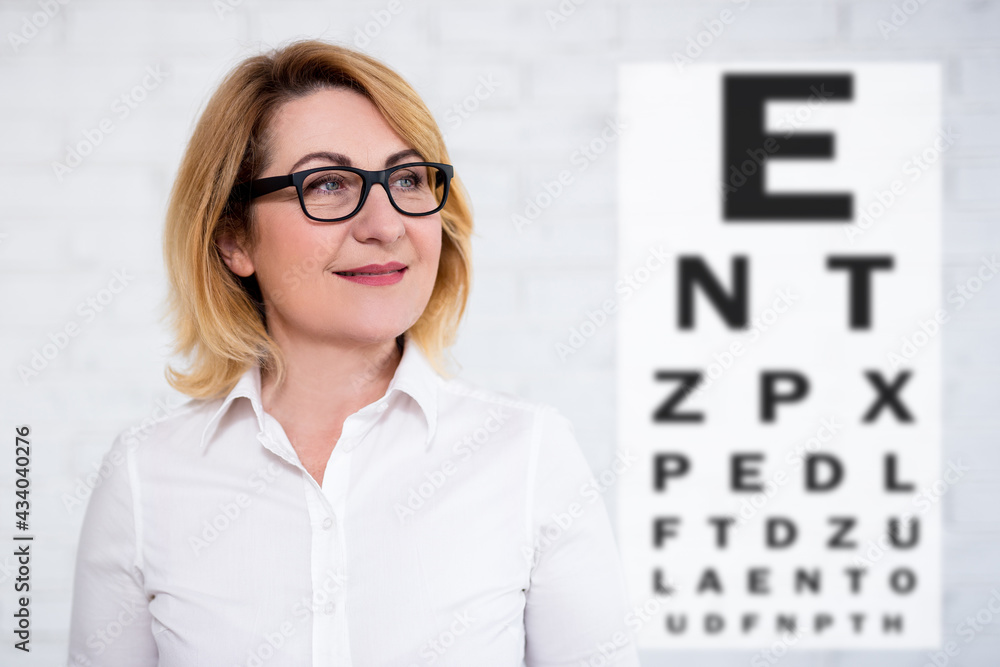 portrait of thoughtful cheerful mature business woman in eyeglasses with eye test chart over white wall portrait of thoughtful cheerful mature business woman in eyeglasses with eye test chart over white wall