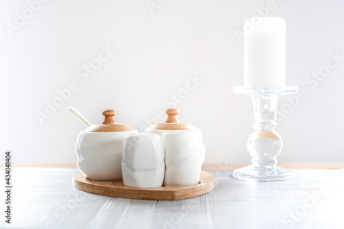 White seasoning containers and a candle on a table. Wooden details on kitchenware set  the stove of the candle made from glass. Selective focus on the small container in front  blurred background.