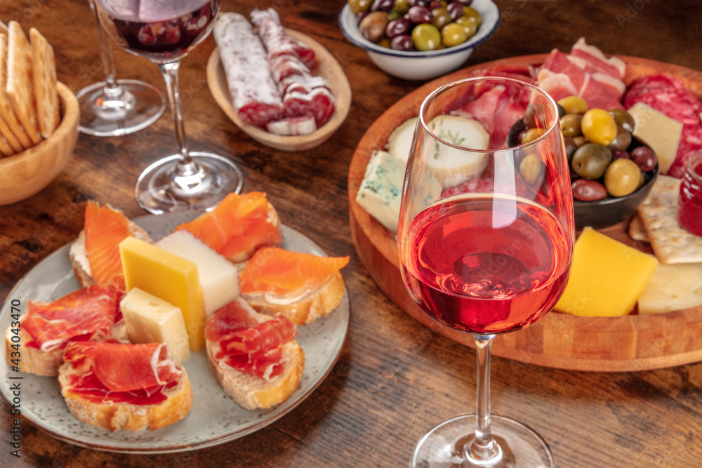 A glass of rose wine with sandwiches and a cheese and charcuterie platter. Italian antipasti or Spanish tapas. Elegant gourmet appetizers on a wooden table A glass of rose wine with sandwiches and a cheese and charcuterie platter. Italian antipasti or Spanish tapas. Elegant gourmet appetizers on a wooden table