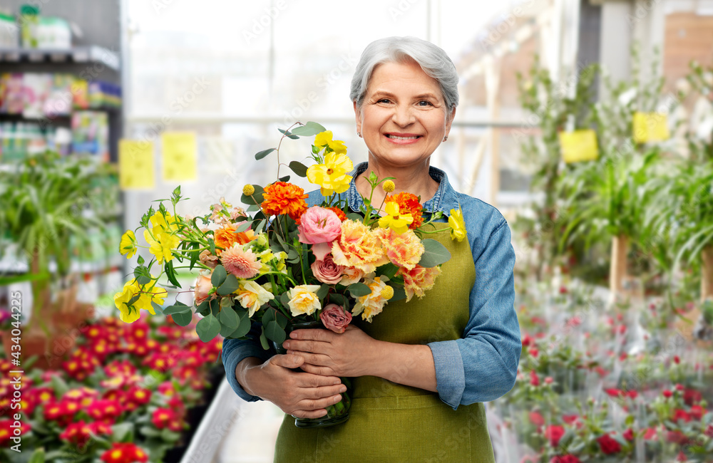 gardening, floristic and old people concept - portrait of smiling senior woman in green apron with bunch of flower over greenhouse at garden store on background