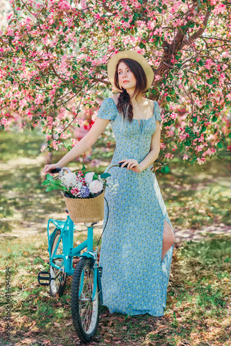 Beautiful girl in blue dress walk with bicycle in blooming spring garden