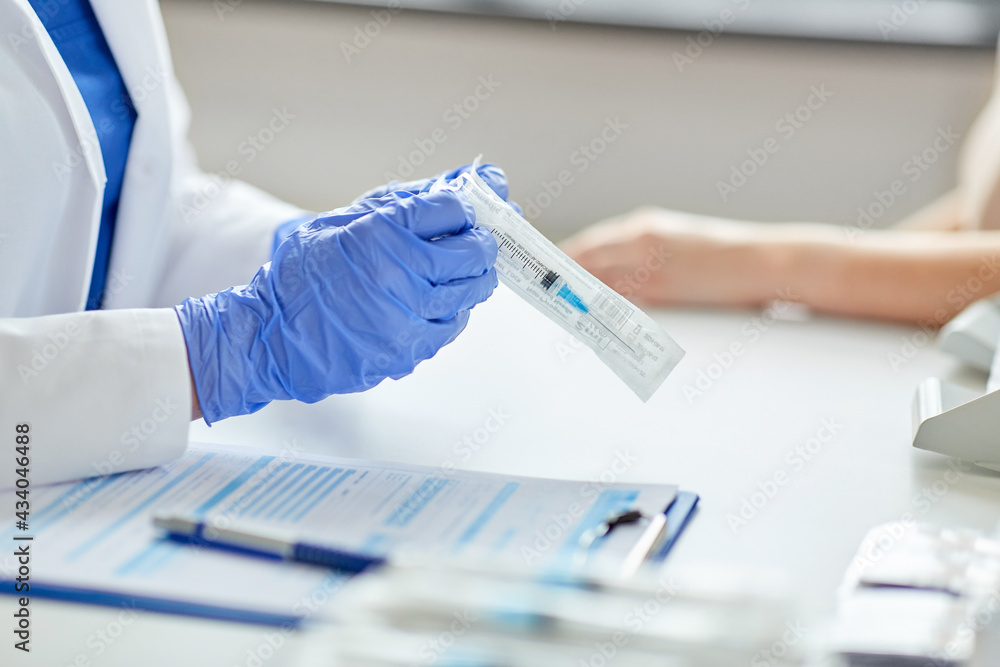 health, medicine and pandemic concept - close up of female doctor or nurse wearing protective medical gloves opening package with syringe for vaccination and patient at hospital health, medicine and pandemic concept - close up of female doctor or nurse wearing protective medical gloves opening package with syringe for vaccination and patient at hospital