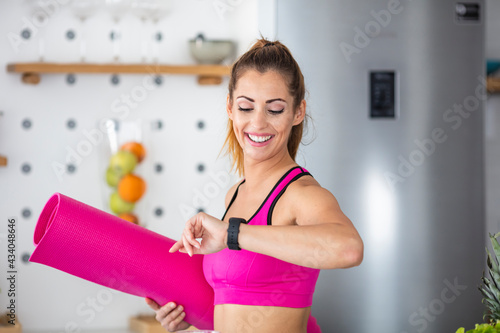 It's time for training. Smiling athletic woman drinking healthy smoothie and checking the time on her wristwatch in the kitchen. She is persistent in her healthy lifestyle!