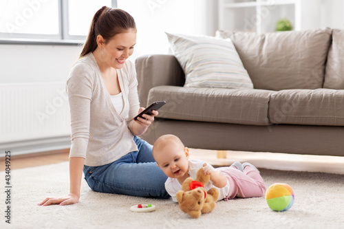 family, motherhood and people concept - happy smiling mother with smartphone and little baby daughter playing with soft ball toy and teddy bear on floor at home