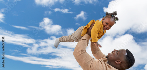family, fatherhood and people concept - happy african american father playing with baby daughter over blue sky and clouds background © Syda Productions