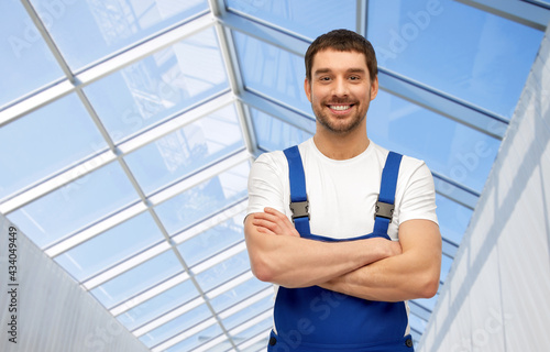 profession, construction and building concept - happy smiling male worker or builder in overall over glasshouse background