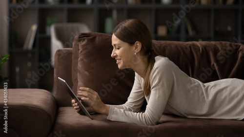 Free time online. Young lady rest with comfort on soft couch scroll news on tablet device using wifi. Happy teen female electronic pad user enjoy video content at social network chat read digital book