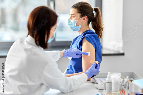 health, medicine and vaccination concept - female doctor wearing protective medical mask and gloves attaching adhesive wound plaster or patch to medical worker at hospital