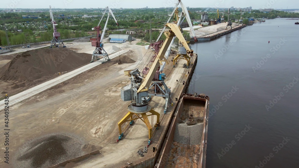 Large harbor cranes onshore at the quay unload a river gravel barge onto a large pile. Cargo port operation. Drone quadrocopter for aerial photography.