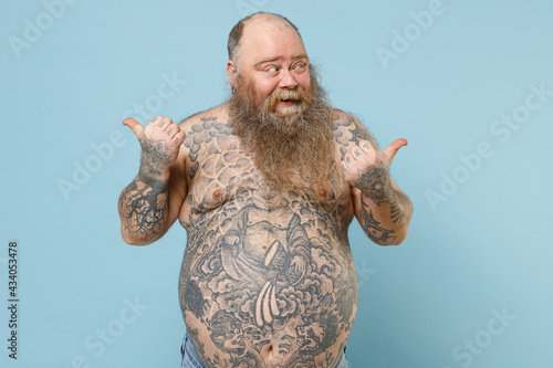 Young fat pudge obese chubby overweight blue-eyed bearded man 30s has big belly with naked tattooed torso point thumb aside back on worksace area isolated on pastel blue background studio portrait photo