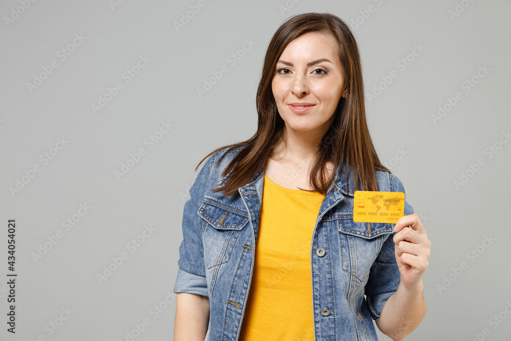 Young smiling happy caucasian woman 20s in casual trendy denim jacket yellow t-shirt holding in hand credit bank card look camera isolated on grey background studio portrait. People lifestyle concept Young smiling happy caucasian woman 20s in casual trendy denim jacket yellow t-shirt holding in hand credit bank card look camera isolated on grey background studio portrait. People lifestyle concept