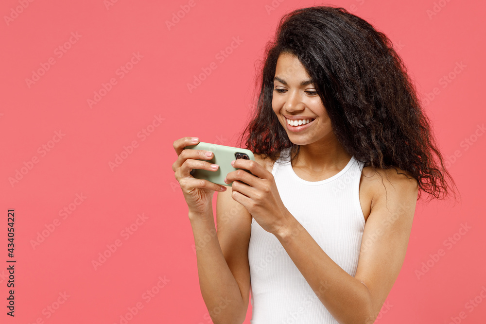 Young gambling african american woman in white tank shirt do selfie shot on mobile phone using play racing on mobile cell phone hold gadget smartphone for pc video games isolated on pink background Young gambling african american woman in white tank shirt do selfie shot on mobile phone using play racing on mobile cell phone hold gadget smartphone for pc video games isolated on pink background
