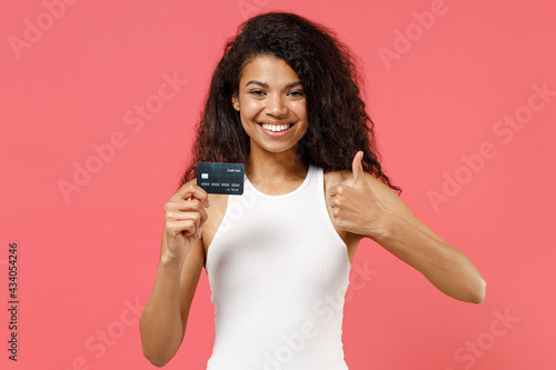 Young fun smiling happy successful satisfied positive african american woman in white tank shirt hold in hand credit bank card show thumb up like gesture isolated on pink background studio portrait.