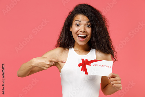 Young excited happy satisfied surprised african american woman 20s wearing casual white tank shirt point index finger on gift voucher flyer mock up isolated on pink color background studio portrait.