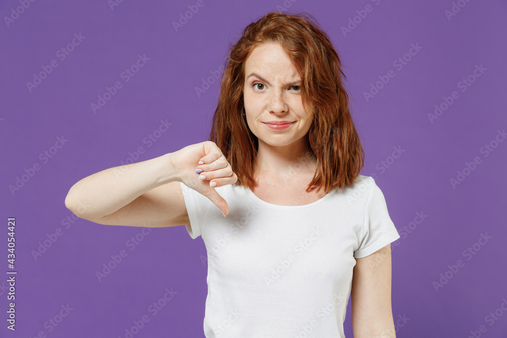 Fototapeta premium Young dissatisfied displeased sad disappointed caucasian woman in white basic blank print design t-shirt showing thumb down dislike gesture isolated on dark violet color background studio portrait