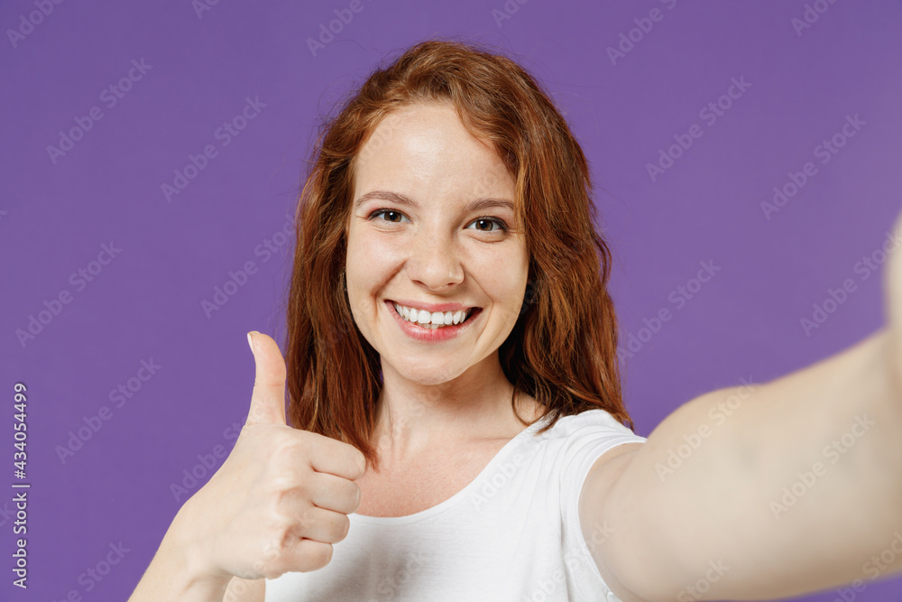 Close up young smiling happy friendly redhead woman 20s wear white basic casual t-shirt doing selfie shot on mobile phone show thumb up like gesture isolated on dark violet color background studio Close up young smiling happy friendly redhead woman 20s wear white basic casual t-shirt doing selfie shot on mobile phone show thumb up like gesture isolated on dark violet color background studio