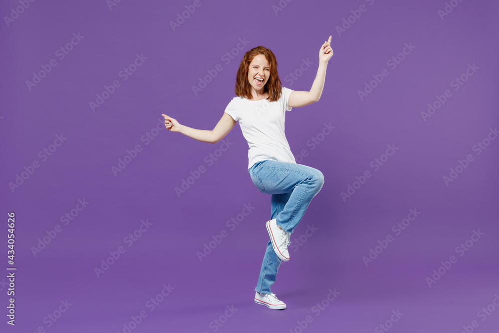 Full length young fun overjoyed redhead caucasian woman 20s in white basic casual t-shirt jeans do winner gesture point index finger up with raised up leg isolated on dark violet background studio