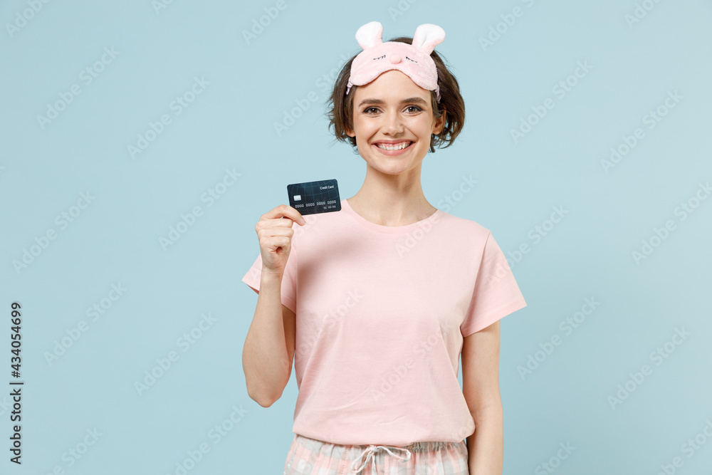 Young smiling successful happy woman 20s wear pajamas jam sleep eye mask rest relaxing at home hold credit bank card isolated on pastel blue color background studio. Good mood night bedtime concept. Young smiling successful happy woman 20s wear pajamas jam sleep eye mask rest relaxing at home hold credit bank card isolated on pastel blue color background studio. Good mood night bedtime concept.