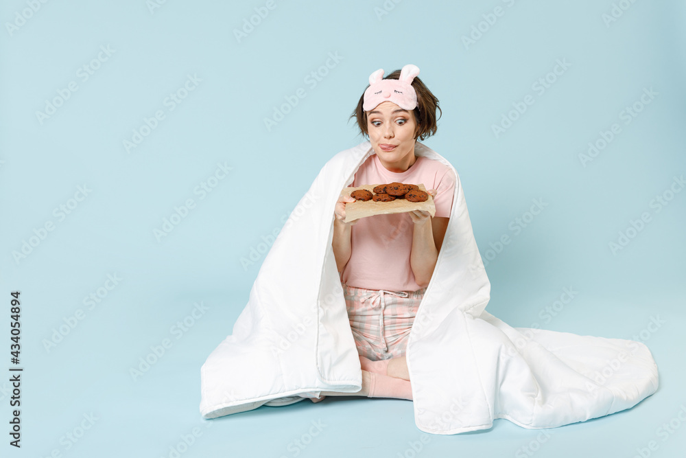 Full length young confused woman 20s in pajamas jam sleep eye mask rest at home wrap cover blanket duvet sit eat pastry sweet cookie eat isolated on pastel blue background Good mood breakfast concept. Full length young confused woman 20s in pajamas jam sleep eye mask rest at home wrap cover blanket duvet sit eat pastry sweet cookie eat isolated on pastel blue background Good mood breakfast concept.