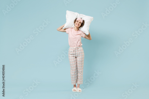 Full length young dreamful happy woman 20s in pajamas jam sleep eye mask rest relax at home holding pillow look aside isolated on pastel blue color background studio. Good mood night bedtime concept