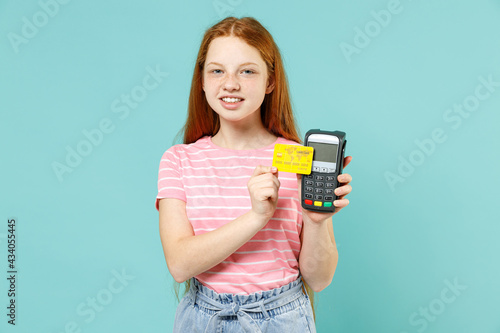 Little fun redhead kid girl 12-13 year old in pink tshirt hold wireless modern bank payment terminal process acquire credit card payments isolated on pastel blue background Lifestyle children concept