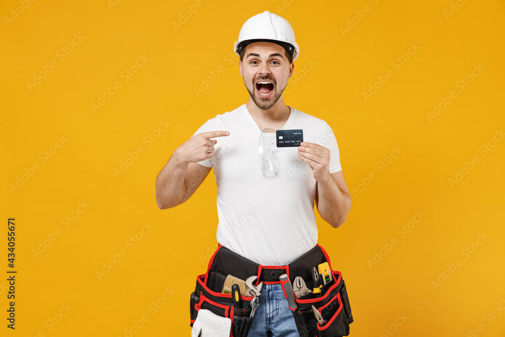 Young employee handyman man in protective helmet hardhat point index finger on credit bank card isolated on yellow background Instruments accessories for renovation apartment room Repair home concept.