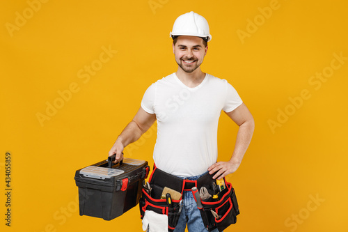 Young smiling excited confident employee handyman man 20s in protective helmet hardhat tool case box isolated on yellow background Instruments accessory renovation apartment room Repair home concept photo