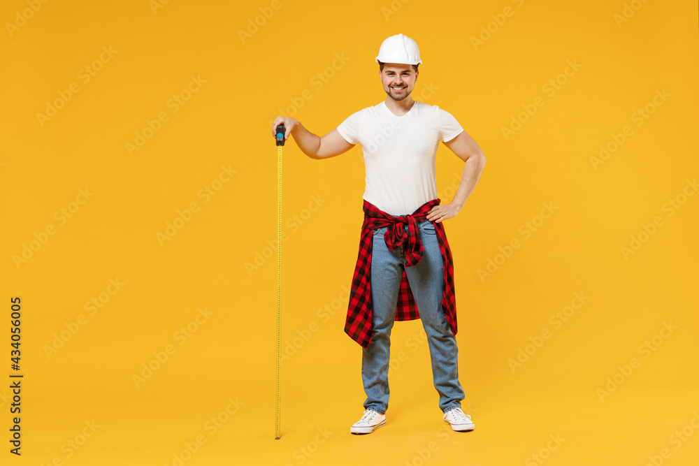 Full length happy young employee handyman man in protective helmet hold measurement tape stand akimbo isolated on yellow background Instrument accessory renovation apartment room Repair home concept Full length happy young employee handyman man in protective helmet hold measurement tape stand akimbo isolated on yellow background Instrument accessory renovation apartment room Repair home concept