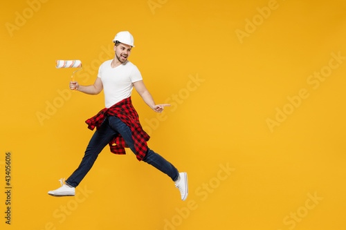 Full length young employee handyman man in t-shirt using paint roller jump high point finger aside isolated on yellow background Instruments accessories renovation apartment room. Repair home concept. © ViDi Studio