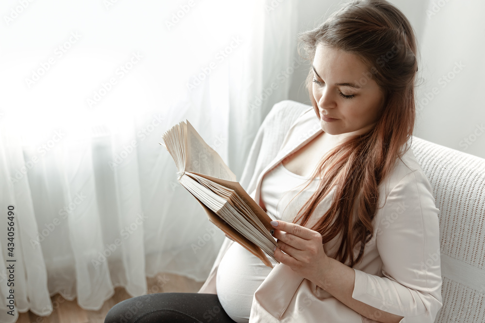 Young pregnant woman reading a book while sitting on the sofa at home. Young pregnant woman reading a book while sitting on the sofa at home.