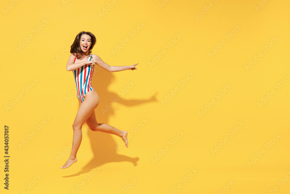 Full length fun happy young sexy woman slim body wear striped red blue one-piece swimsuit jump high air run fly isolated on vivid yellow color background studio Summer hotel pool sea rest sun concept