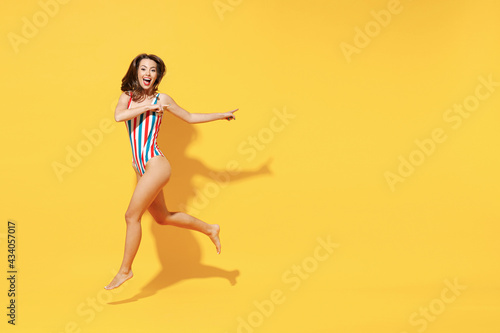 Full length fun happy young sexy woman slim body wear striped red blue one-piece swimsuit jump high air run fly isolated on vivid yellow color background studio Summer hotel pool sea rest sun concept