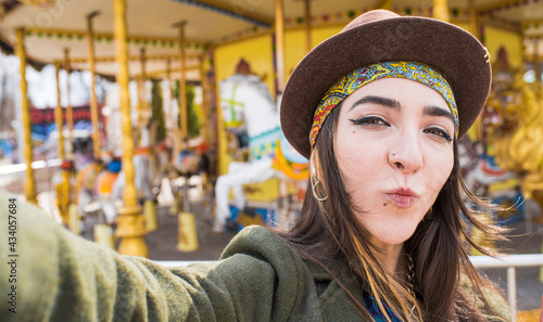 Portrait of a hippie girl in a hat who takes a selfie on a smartphone and shows a kiss in the park near the attraction
