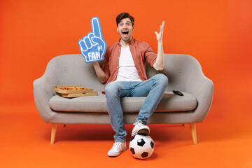 Young surprise man football fan in shirt support team with soccer ball sit on home sofa watch tv live stream hold pizza foam glove finger spread arm isolated on orange background People sport concept